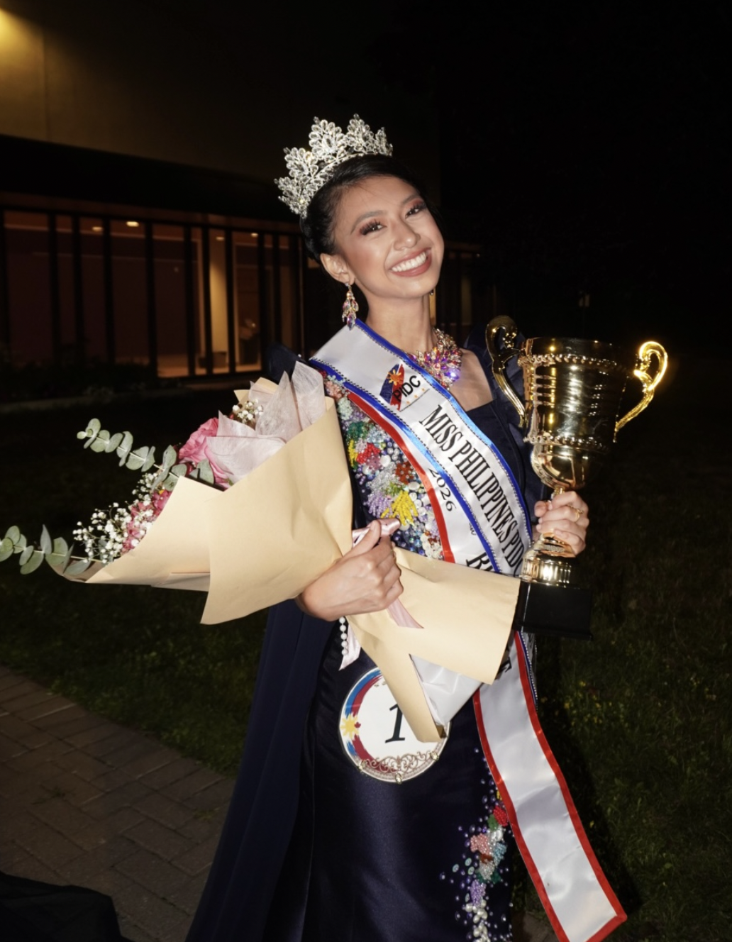 Isabella wearing her pageant sash and crown, holding flowers and a trophy